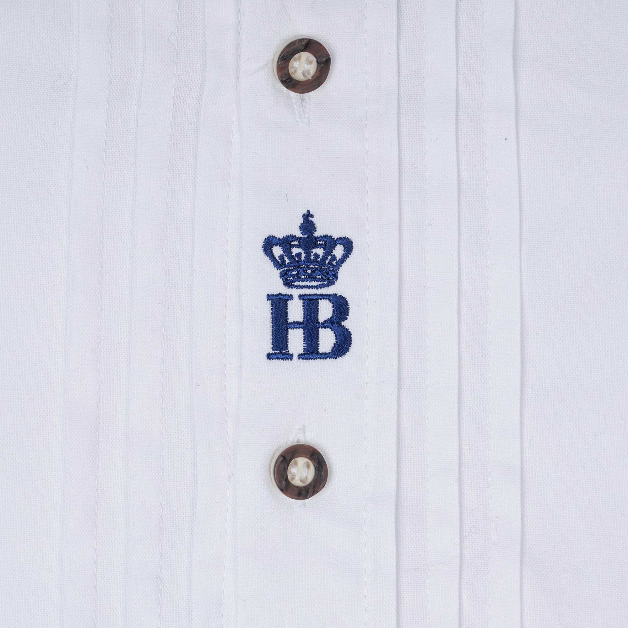 Nahaufnahme eines weißen Hemds mit dunkelblauer „HB“-Stickerei unter einer Krone und braunen Knöpfen.
