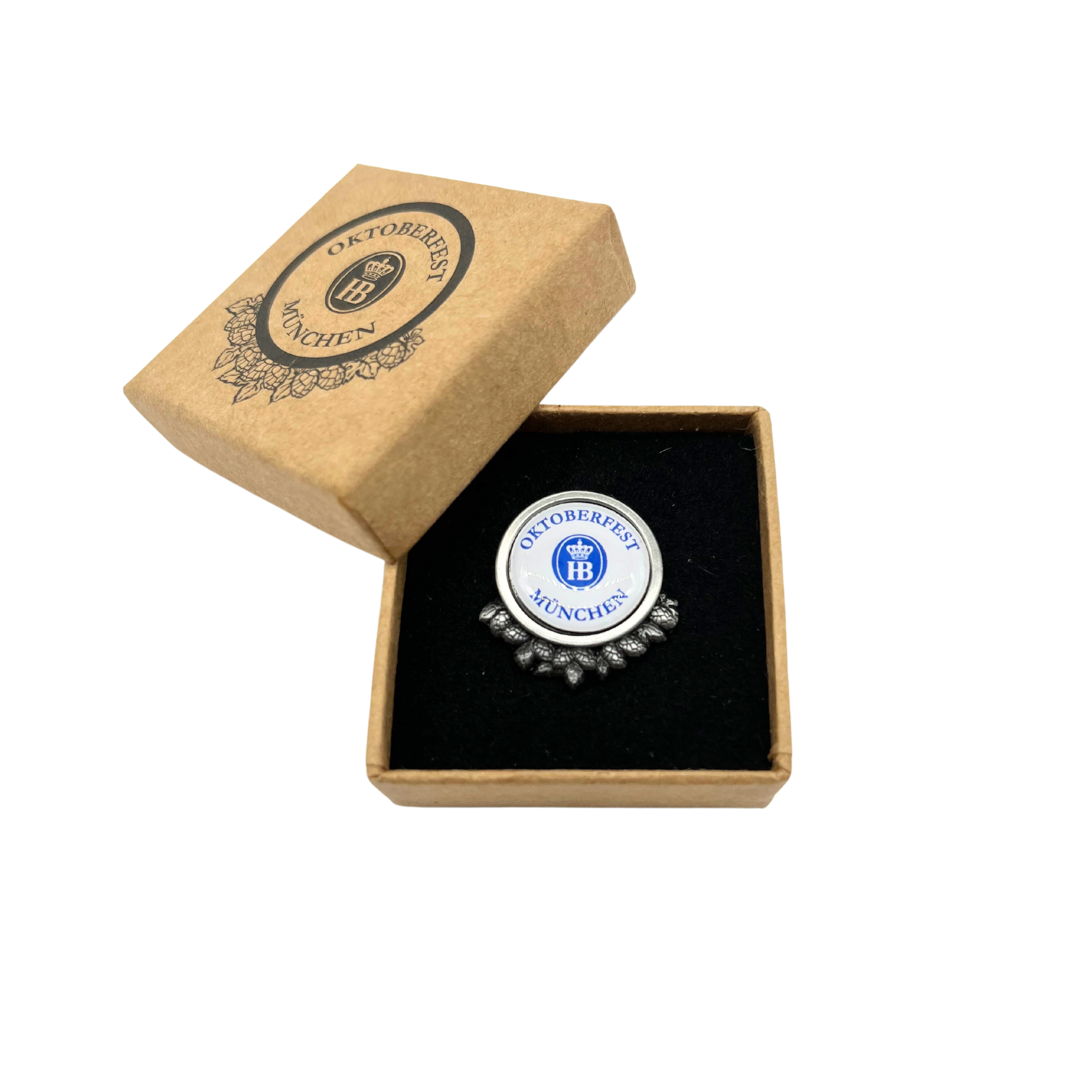 „Quadratische, hellbraune Geschenkbox aus Karton mit schwarzem Innenfutter. Der Deckel ist teilweise geöffnet und zeigt ein rundes, verziertes Design mit der Aufschrift ‚Oktoberfest München‘ mit dem HB Logo. Im Inneren befindet sich der Pin Oktoberfest mi
