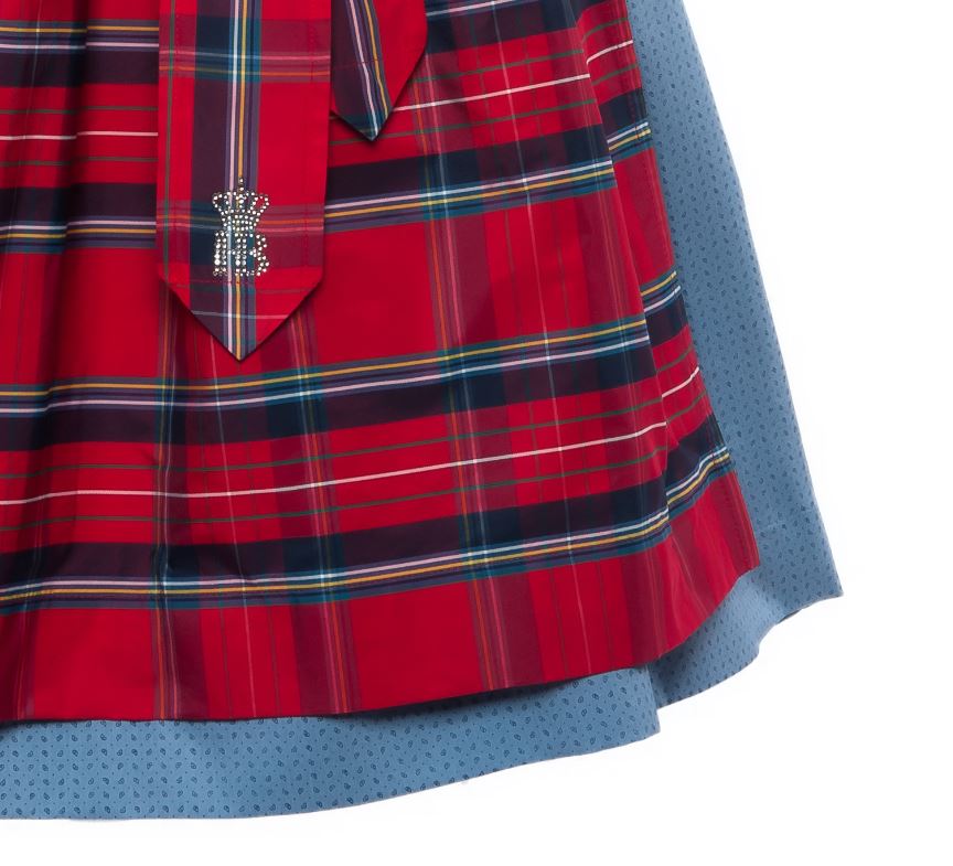 Detailaufnahme eines Dirndl-Schurzes im roten Karomuster mit blauem Unterrock; am Schürzenband glitzerndes Hofbräu-Logo aus Strasssteinen.