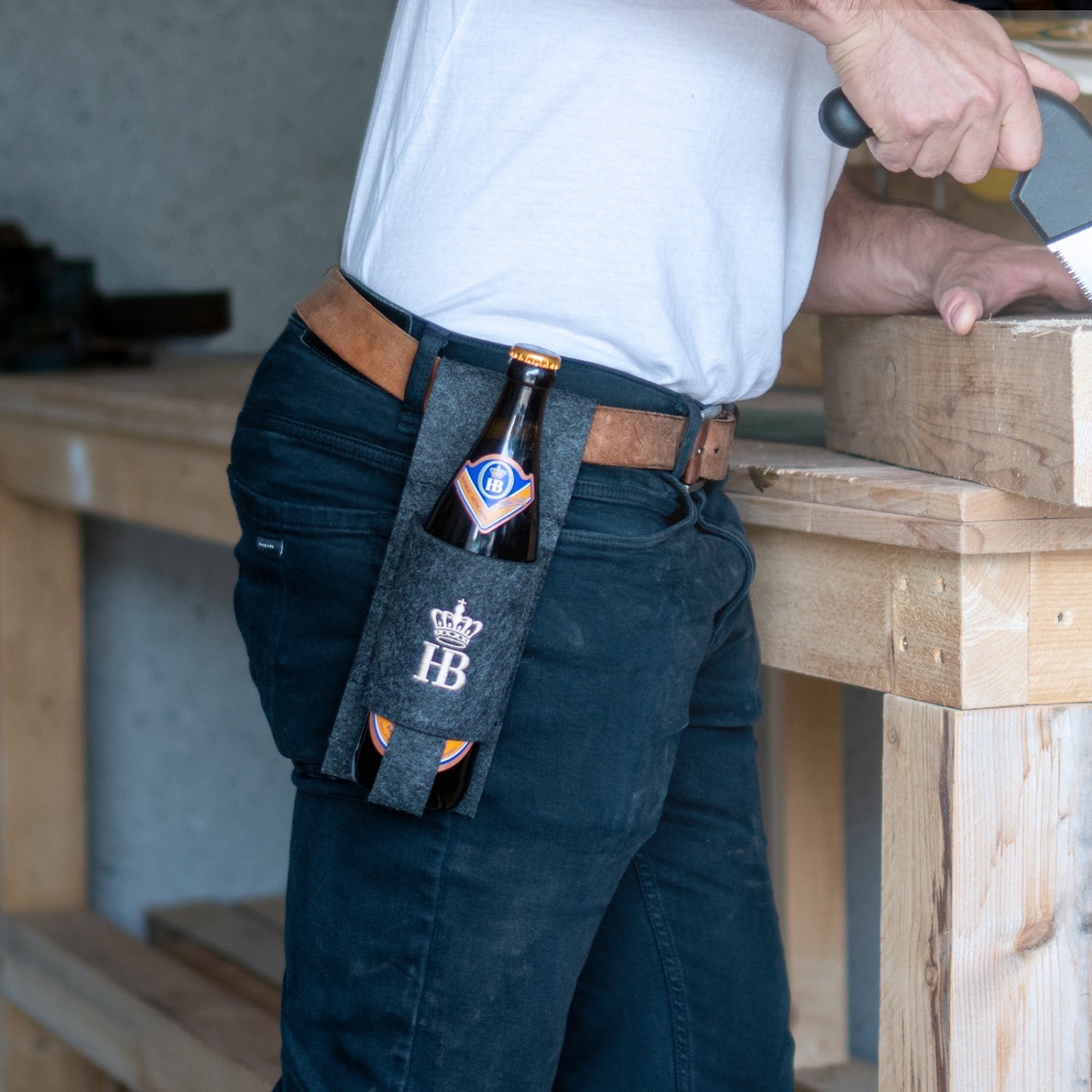 Mann mit weißem T-Shirt und Jeans arbeitet an einer Werkbank, am Gürtel hängt ein dunkler Filz-Flaschenhalter mit HB-Logo, in dem eine Hofbräu-Bierflasche steckt