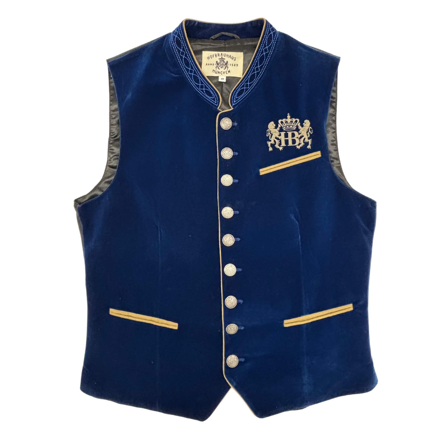 „Blaue Weste mit goldfarbenen Knöpfen und goldverzierten Taschen. Auf der linken Brustseite befindet sich ein gesticktes Emblem mit dem HB-Logo, das von zwei bayerischen Löwen flankiert wird – ein traditionelles Symbol für das Hofbräuhaus München.“
