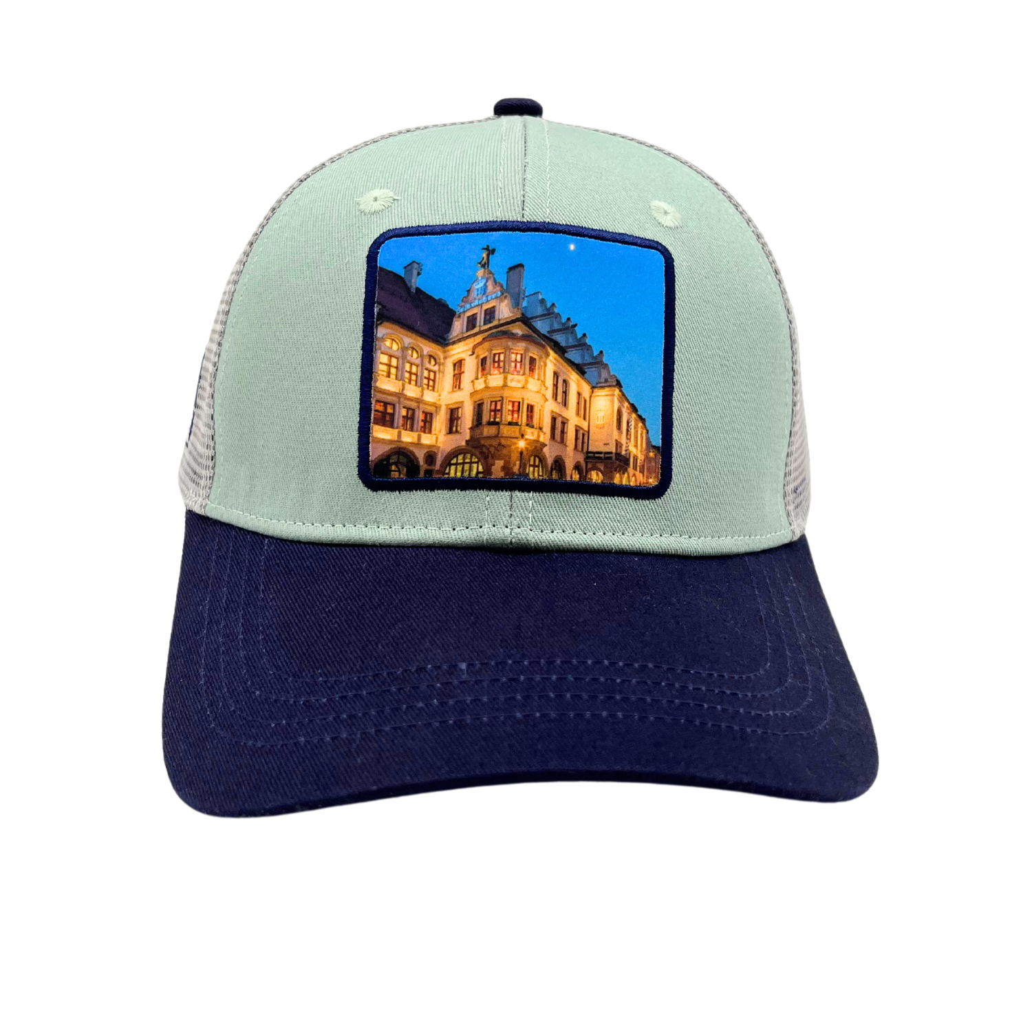 Zweifarbige Trucker-Cap mit hellgrünem Frontpanel, dunkelblauem Schirm und aufgenähtem Patch mit beleuchtetem Hofbräuhaus-Motiv bei Nacht.